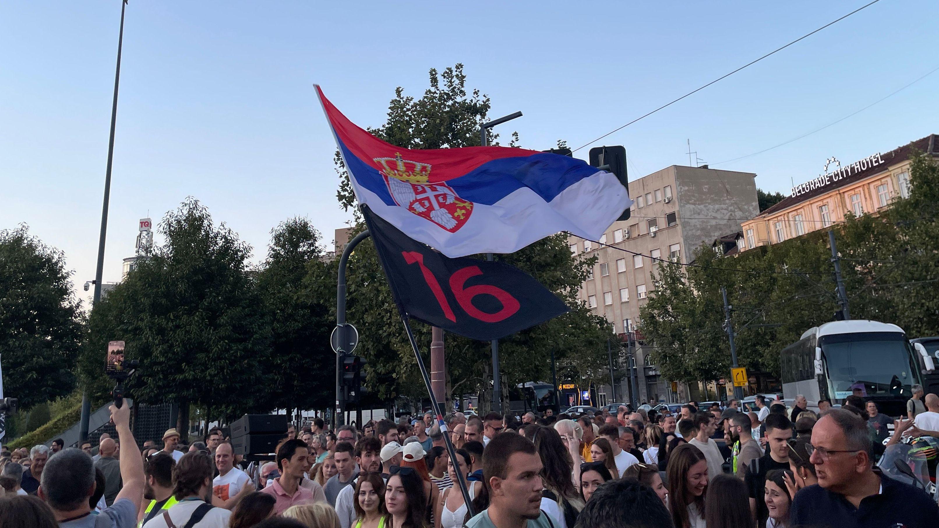 Ljudi na skupu, mađu zastavom srbije i crnom zastavom sa crvenim brojem 16