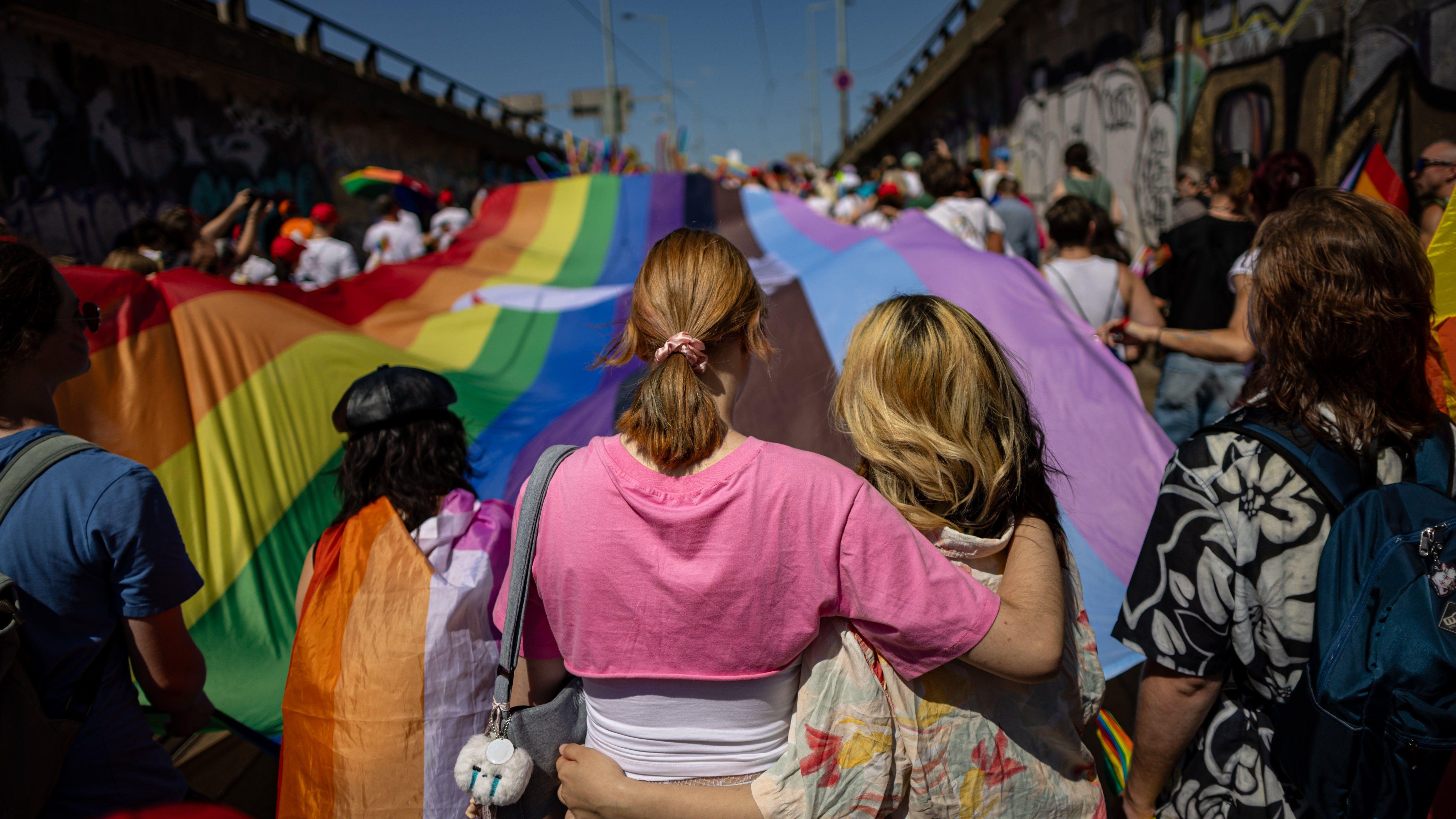 dve devojke zagrljene šetaju u povorci, ispred njih je zastava u duginim bojama 