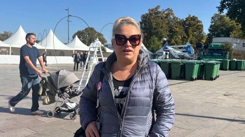 Žena u zimskoj jakni, iza nje muškarac gura kolica, a u daljini su zelene kante za smeće